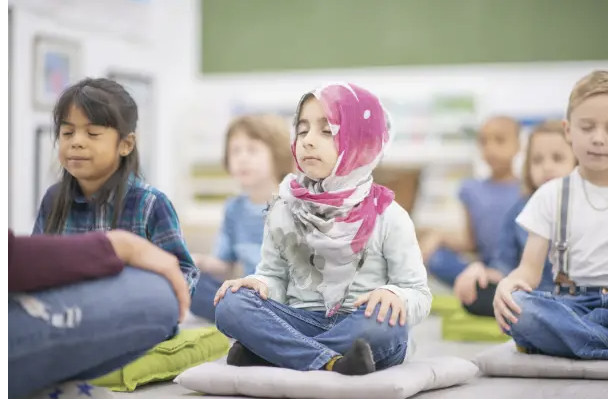 Children sit cross-legged on cushions in a classroom, eyes closed, as if meditating.